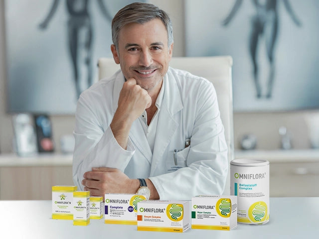 Ein lächelnder männlicher Arzt im weißen Kittel sitzt an einem Schreibtisch, vor dem verschiedene Omniflora-Präparateschachteln und -behälter ausgestellt sind. Im Hintergrund sind unscharfe medizinische Plakate zu sehen.