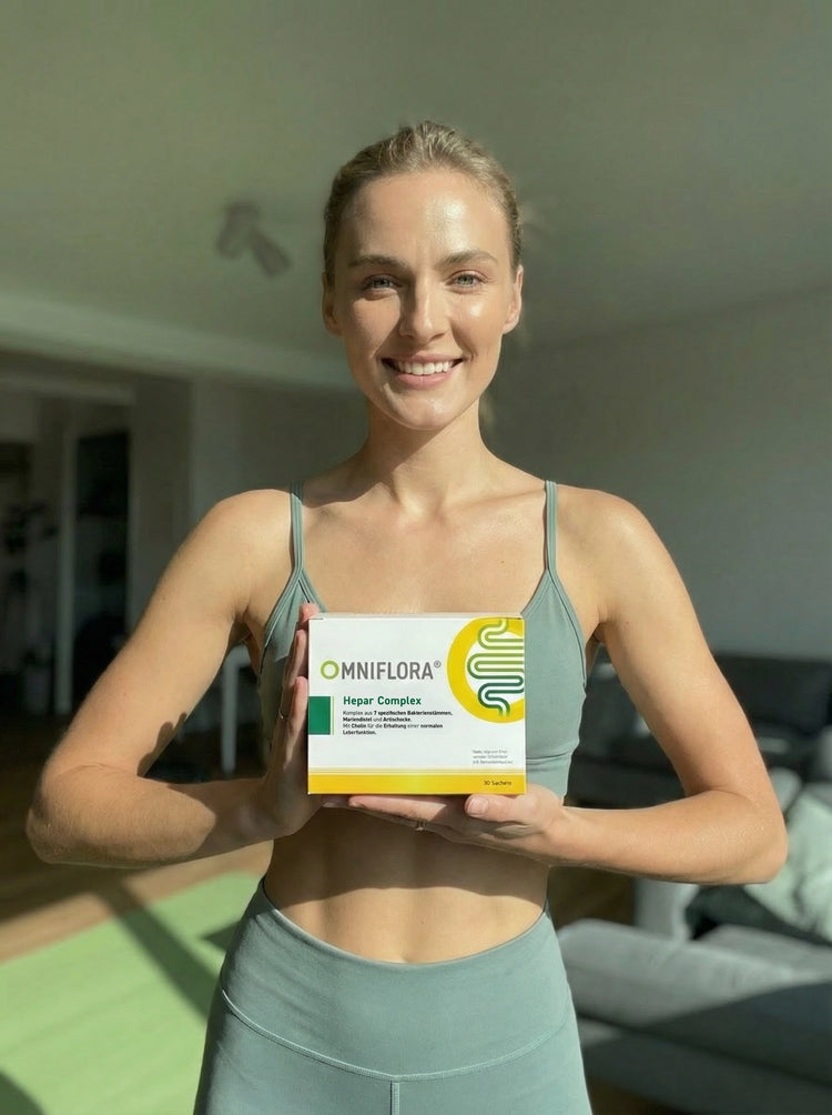 Eine Frau in Sportkleidung steht in einem Innenraum, lächelt und hält eine Schachtel mit der Aufschrift Omniflora+ Hepar Complex in der Hand, in die das Sonnenlicht von der Seite einfällt.