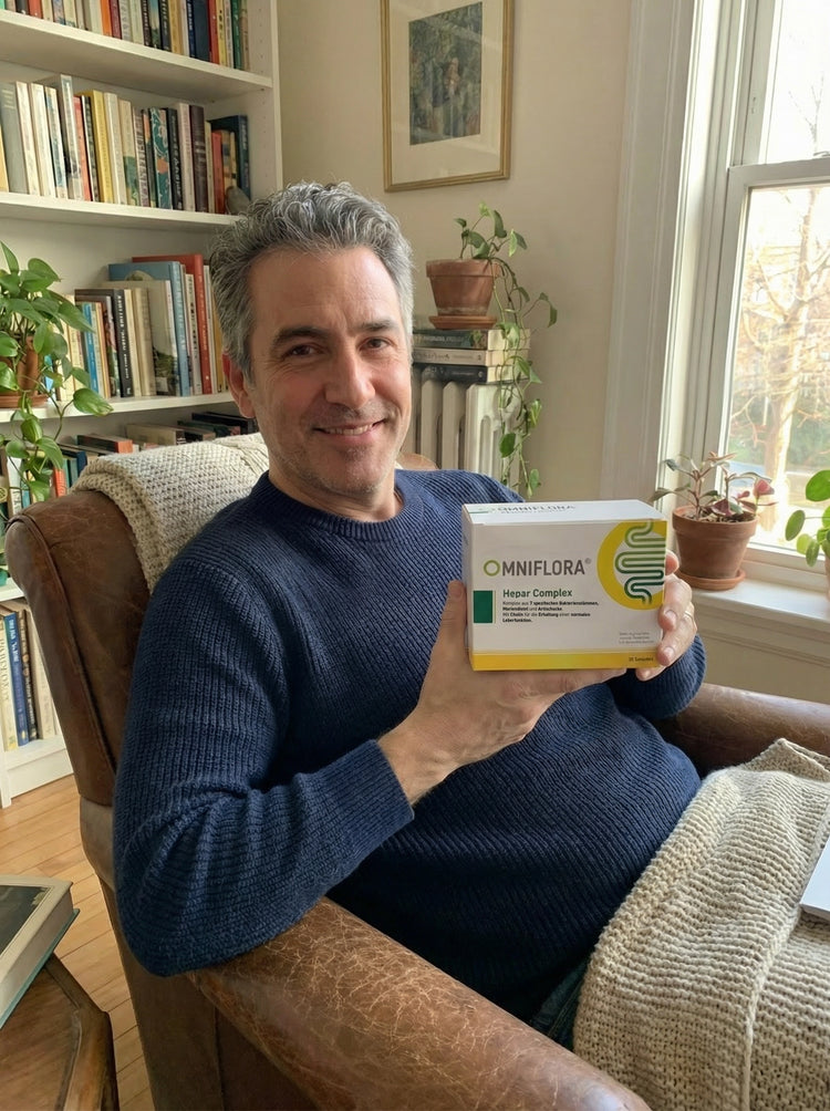 Ein lächelnder Mann mit grauem Haar, der einen marineblauen Pullover trägt, sitzt in einem gemütlichen Sessel und hält eine Schachtel mit der Aufschrift Omniflora Hepat Complex. Er befindet sich in einem Raum mit Bücherregalen, Pflanzen und einem Fenster, das natürliches Licht hereinlässt.
