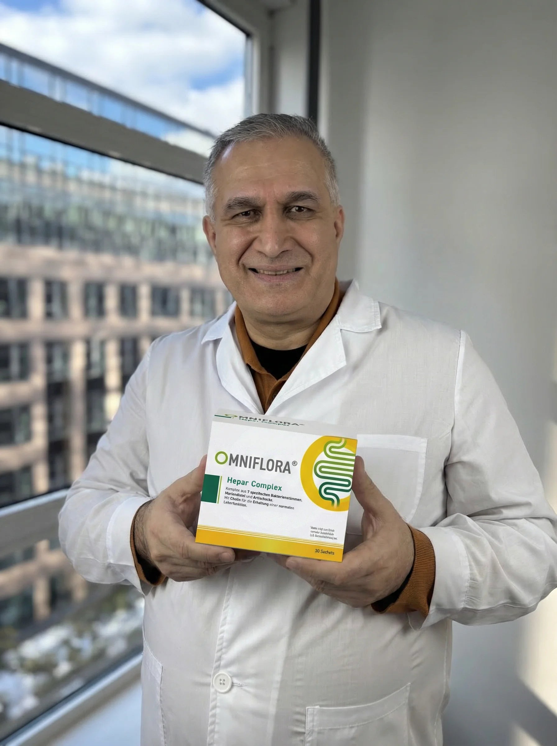Ein lächelnder Mann in einem weißen Laborkittel steht an einem Fenster und hält eine Schachtel mit Omniflora Hepur Complex Ergänzungsmitteln in der Hand. Im Hintergrund ist ein Bürogebäude außerhalb des Fensters zu sehen.